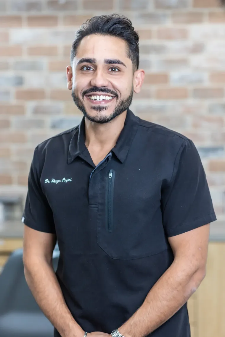 Dr. Shayan Azimi in black scrubs, standing against the exposed brick wall of the Arts Family Dentistry office