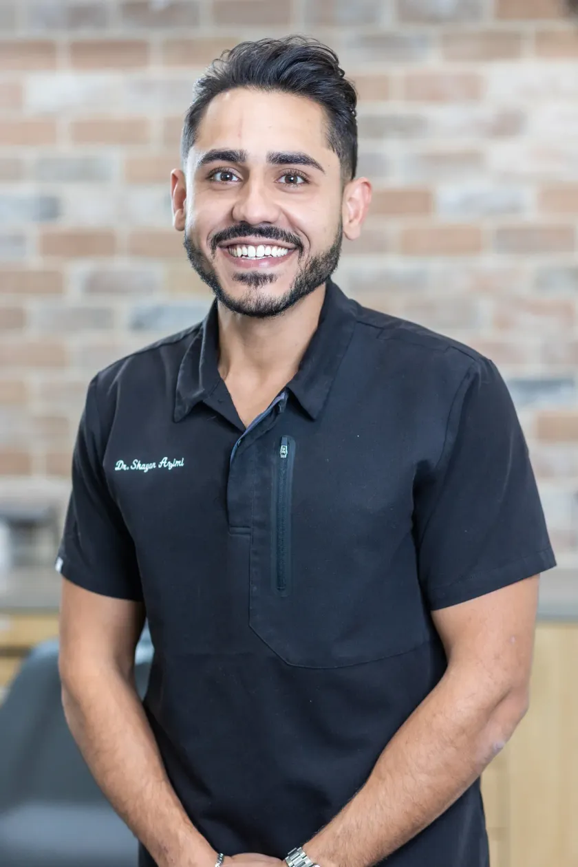 Dr. Shayan Azimi in black scrubs, standing against the exposed brick wall of the Arts Family Dentistry office